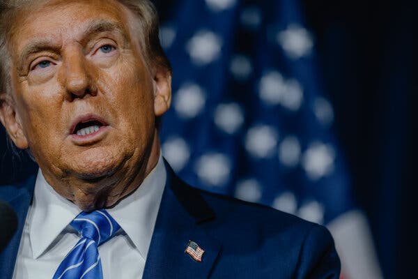 Former President Donald J. Trump wears a blue tie and blue jacket with an American flag pin on the lapel as he speaks during a media appearance.