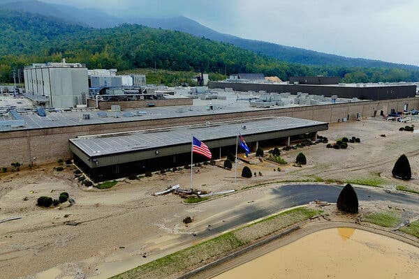 An aerial view of a large facility inundated with mud and brown floodwaters on a rainy day at the foot of a mountain.