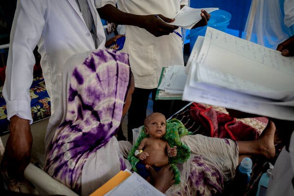 A 3-month-old baby is held by her mother while several doctors in white gowns holding notebooks surround her.