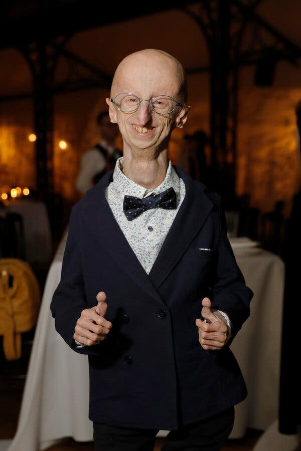 Sammy Basso, wearing a dark double-breasted jacket, a patterned shirt and a bow tie, smiles and gives two thumbs up as he poses for a portrait.