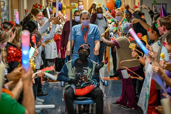 A woman in blue scrubs pushes a boy in a mask and a hat sitting in a wheelchair down a hallway where people on both sides hold light sticks and noisemakers.