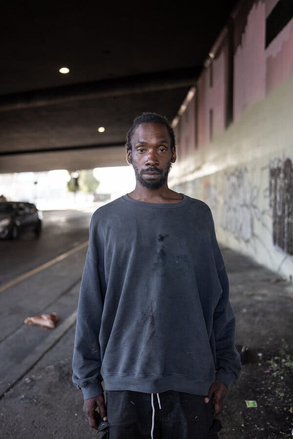 A man in a dark-colored sweatshirt with stains on the front stands under an overpass, looking into the camera.