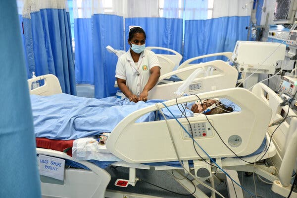 A nurse in a mask stands over a patient hooked up to tubes and wires in a curtained area of a hospital ward. A piece of paper on the bed's side reads "heat stroke patient."