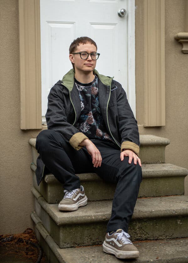 A portrait of Kinnon MacKinnon, who sits on some stone steps leading up to a white door. He wears skateboarder shoes, black pants and a jacket.