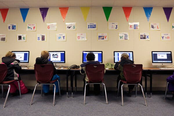 A row of students sitting at computers in a school. A multicolor triangle flag banner is hanging from the wall, and there are small posters with individual alphabet letters on them.