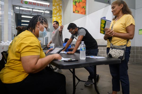 A folding table in the hallway of a food bank has people lined up with documents and computer tablets as they discuss Medicaid enrollment matters.