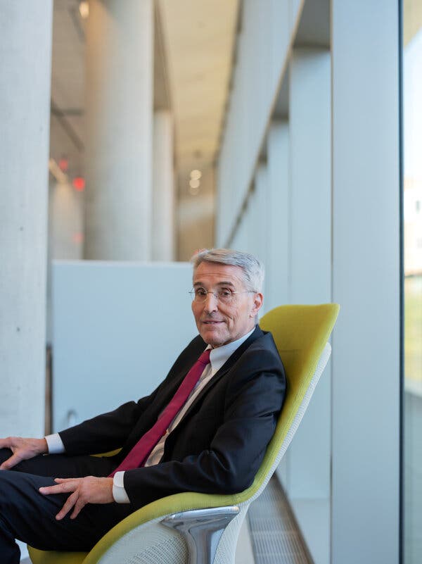 Richard Pazdur, wearing a red necktie and a suit, sits in a yellow chair in a hallway.