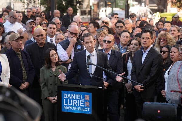 Daniel Lurie speaking from behind a lectern with his arms outstretched and a large crowd behind him. On the lectern are the words “Daniel Lurie, mayor-elect.”