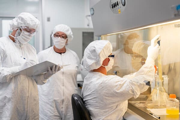 A scientist in a lab facility seated at a workstation manipulates liquids in vials and glassware while two scientists standing behind look at something in a binder. They all wear white suits, hair coverings, face masks and eyewear.