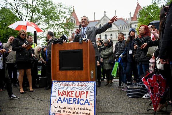 Robert F. Kennedy Jr. speaks from a lectern with a microphone in his hand, gesturing with his other hand. He stands before a crowd at the New York State Capitol and a hand-made sign at the base of the lectern reads "Wake up! America, we have sickest children in all the developed world."