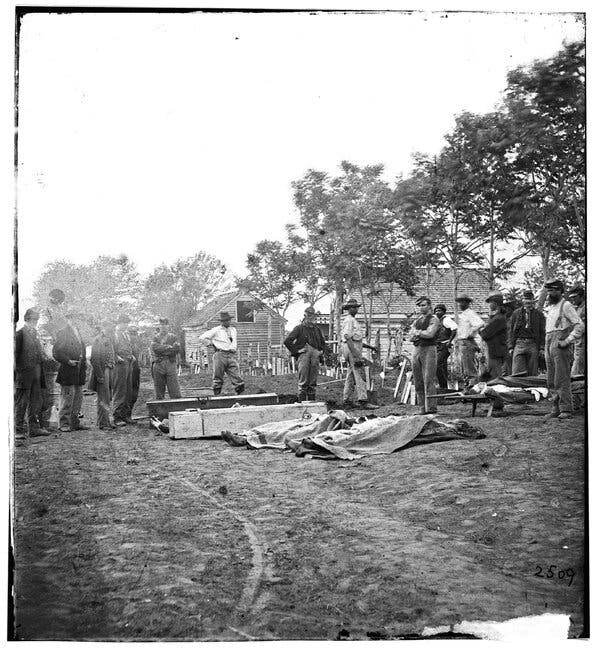 A black-and-white archival photograph of a group of men standing around a lineup of covered bodies ready for burial next to a few wooden coffins.