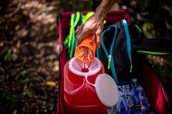 A person’s hand dumping an orange cup of used needles into a red safety disposal bucket in a woodsy area. The bucket is in a cart with other supplies.