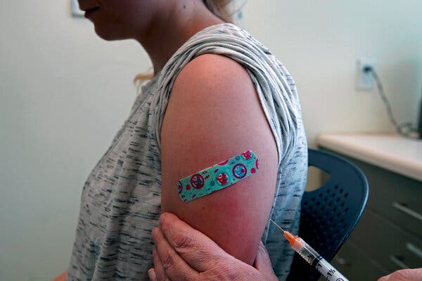 A view of a young adult receiving a vaccine in their left arm. The sleeve is rolled up and there is a bandage on it already from a previous inoculation.