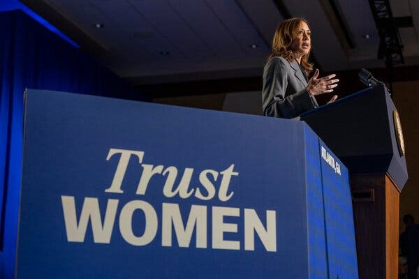 Kamala Harris stands at a podium with a large blue banner that says Trust Women.