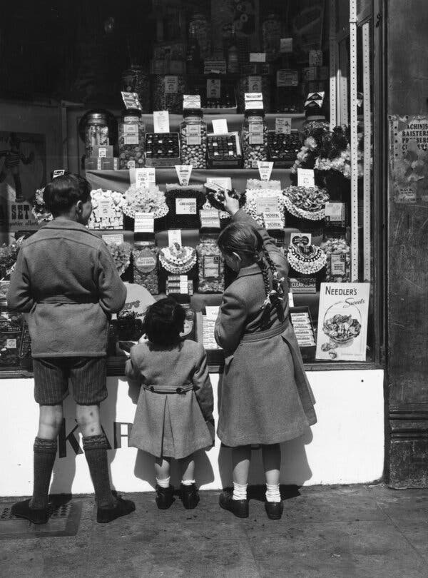 A black and white photograph of three small children lined up outside the window of a candy shop.