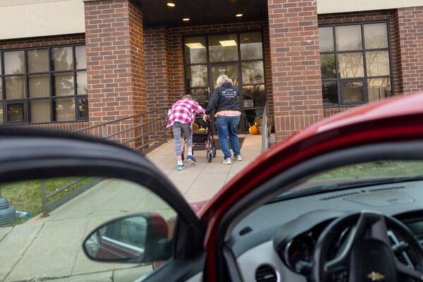 Two people, one behind a walker and one next to it, head toward the glass door of a brick building. The open door of a car is in the foreground.