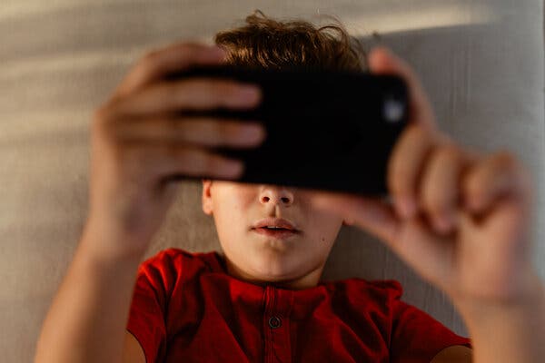 A boy lying on a beige surface watches videos on a mobile phone.