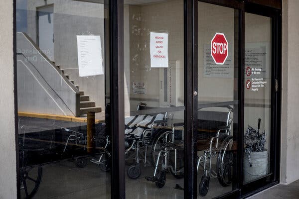 A closed glass door of a hospital with a stop sign sticker and note saying the hospital is closed in red letters, with wheelchairs lined up just behind the door.