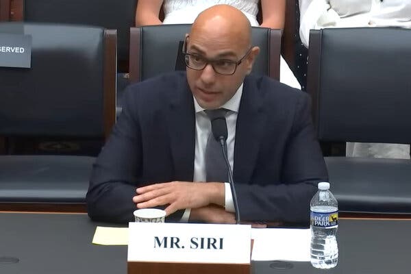 A screengrab from a video of Aaron Siri, who wears a dark blue suit and gray tie, speaking at a microphone at a table in front of a government committee out of view.