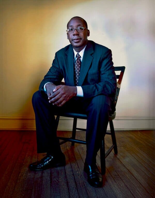 Cornelius Baker sits in a chair, wearing a dark suit and shoes, a white shirt and a polka-dot tie.