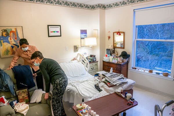 A person helps a nursing home resident rearrange a chair in their room in winter, with snow falling outside the window.