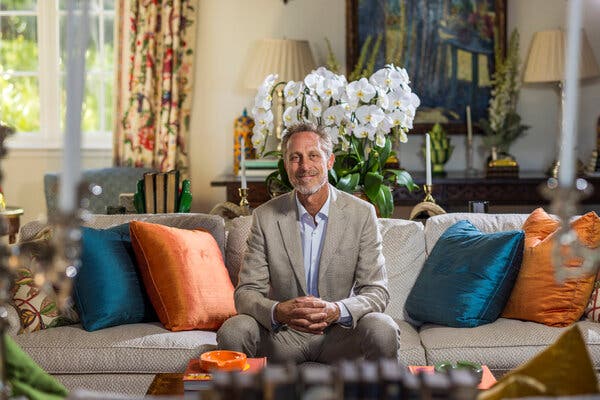 A man in a gray suit sits on a couch with blue and orange pillows by his side.