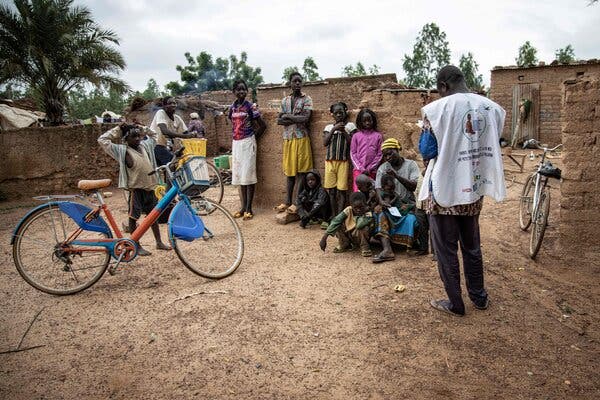 A half-dozen children and teenagers stand on a dirt expanse leaning against a low wall. Some have bicycles parked. A man with his back to the camera is addressing them.