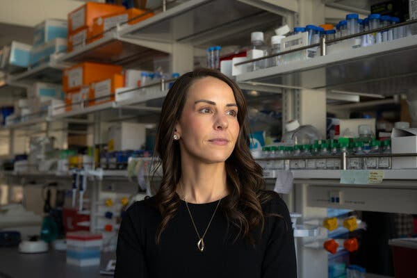 A woman wearing a black shirt and silver necklace stands in front of laboratory shelves looking to the side.