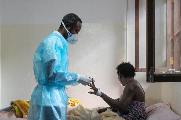 A man in a surgical mask, gown and gloves stands over a patient in a bed, who is looking away.