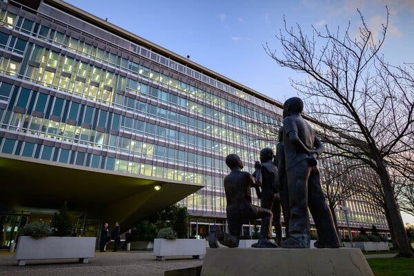 The exterior of the World Health Organization headquarters in Geneva on a winter day. In front of the building is a sculpture of a figure kneeling to administer a vaccine to a child.