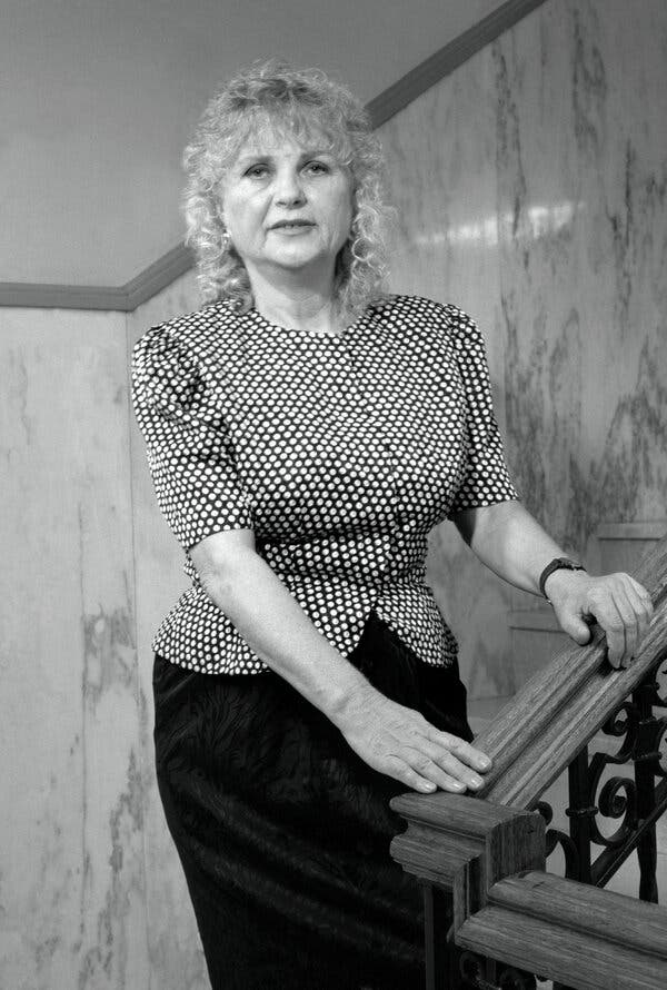 A black-and-white photo of Carol Downer, a woman with curly shoulder-length blond hair, standing in a stairway with both hands on the railing.