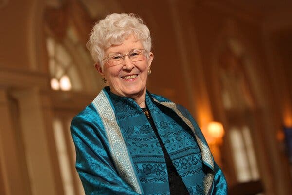 A portrait of her smiling at the camera in an elegant-looking room. She has short, curly white hair and wears eyeglasses and a blue-patterned outfit.