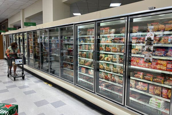 00food-safety-cuts-01-lqpm-articlelarge A woman pushing a grocery cart along an aisle of refrigerated glass-door cabinets of foods.