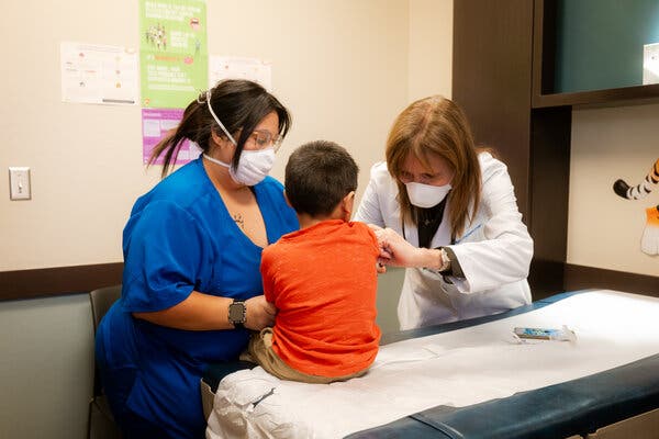 00measles-01b-mlqz-articlelarge A nurse practitioner wearing a white mask gives a shot to a boy sitting on an exam room table, as another medical professional holds the boy’s other arm.