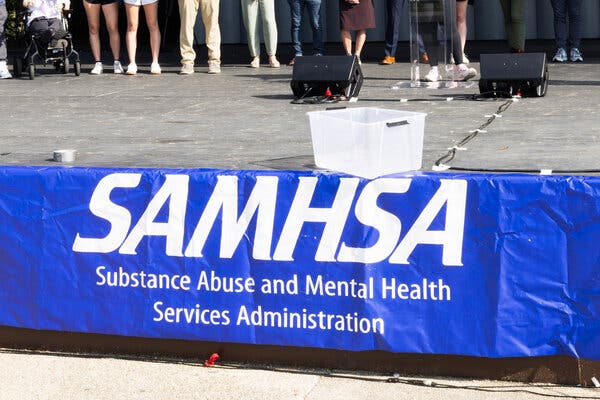 12addiction-cuts-articlelarge A big blue banner reads “SAMHSA” and “Substance Abuse and Mental Health Services Administration” on it in white lettering. The legs of people standing behind it are visible.