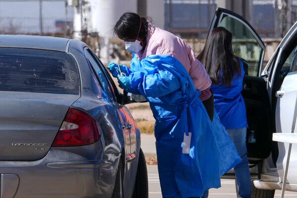 14measles-01-fjkp-articlelarge A person in a mask and blue P.P.E. and gloves stands at the passenger window of a car to administer a measles swab test.
